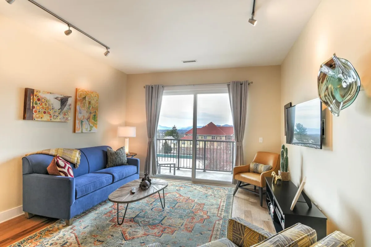 A stylish living area with a sofa and coffee table arranged to face a large window, which offers a glimpse of the vibrant cityscape outside. Colorful artwork decorates the walls, adding an artistic touch to the comfortable, inviting space. Morning light spills in, while the eclectic decor nods to the creative energy of the neighborhood.