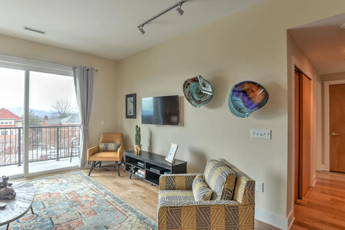 This interior photo captures a comfortable living area with a clear view through the balcony, colorful wall decor, and inviting furnishings. The visible balcony and cityscape hint at direct access to the outdoors and the urban landscape beyond.
