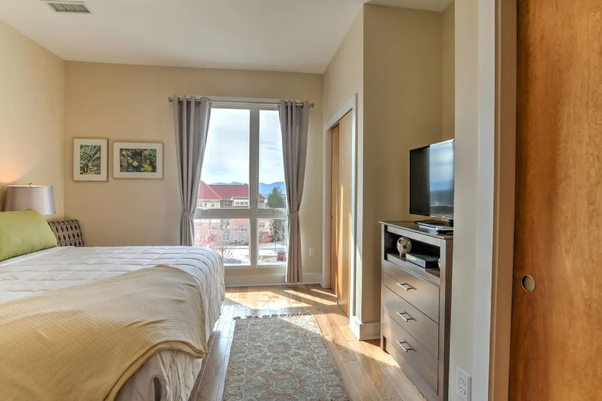 A serene bedroom with a king bed draped in a textured blanket, illuminated by sunlight streaming through a large window. Framed artwork and a television unit add modern touches, creating a relaxing, contemporary space ideal for recharging.