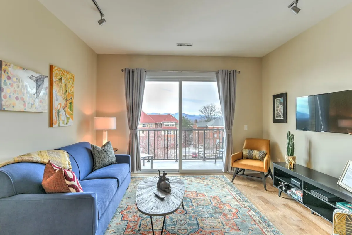 A cozy living room setup featuring a blue sofa, artistic wall decor, and a large window offering a glimpse of the city outside. The scene radiates warmth and showcases how residents can admire Asheville from the comfort of home.