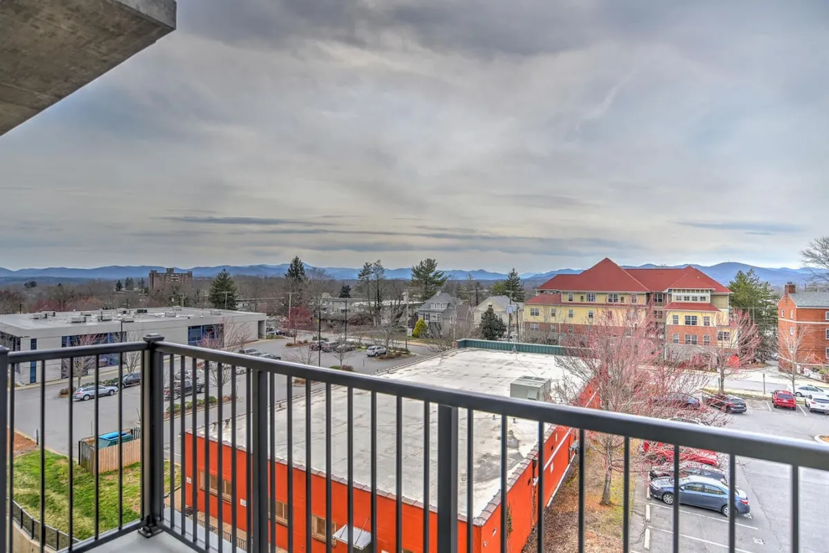 This image presents a sweeping view of Asheville’s urban skyline against a backdrop of mountains, with a colorful building adding vibrancy to the cityscape. The vantage point likely comes from a balcony or rooftop, evoking a sense of place and immediate access to the city’s energy.