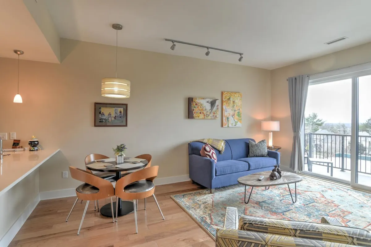 This image captures an open-plan living and dining area with a blue sofa, a modern dining table, and art adorning the walls. The space is bathed in natural light, and the balcony doors reveal inviting outdoor views, seamlessly blending the interior with the exterior.