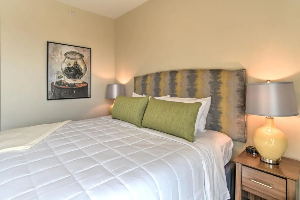 A cozy king-sized bed topped with decorative pillows and a soft comforter sits under a piece of elegant artwork, with a nightstand and lamp adding warmth and convenience. The overall ambiance is calm and serene, perfect for unwinding after a day of city adventures.