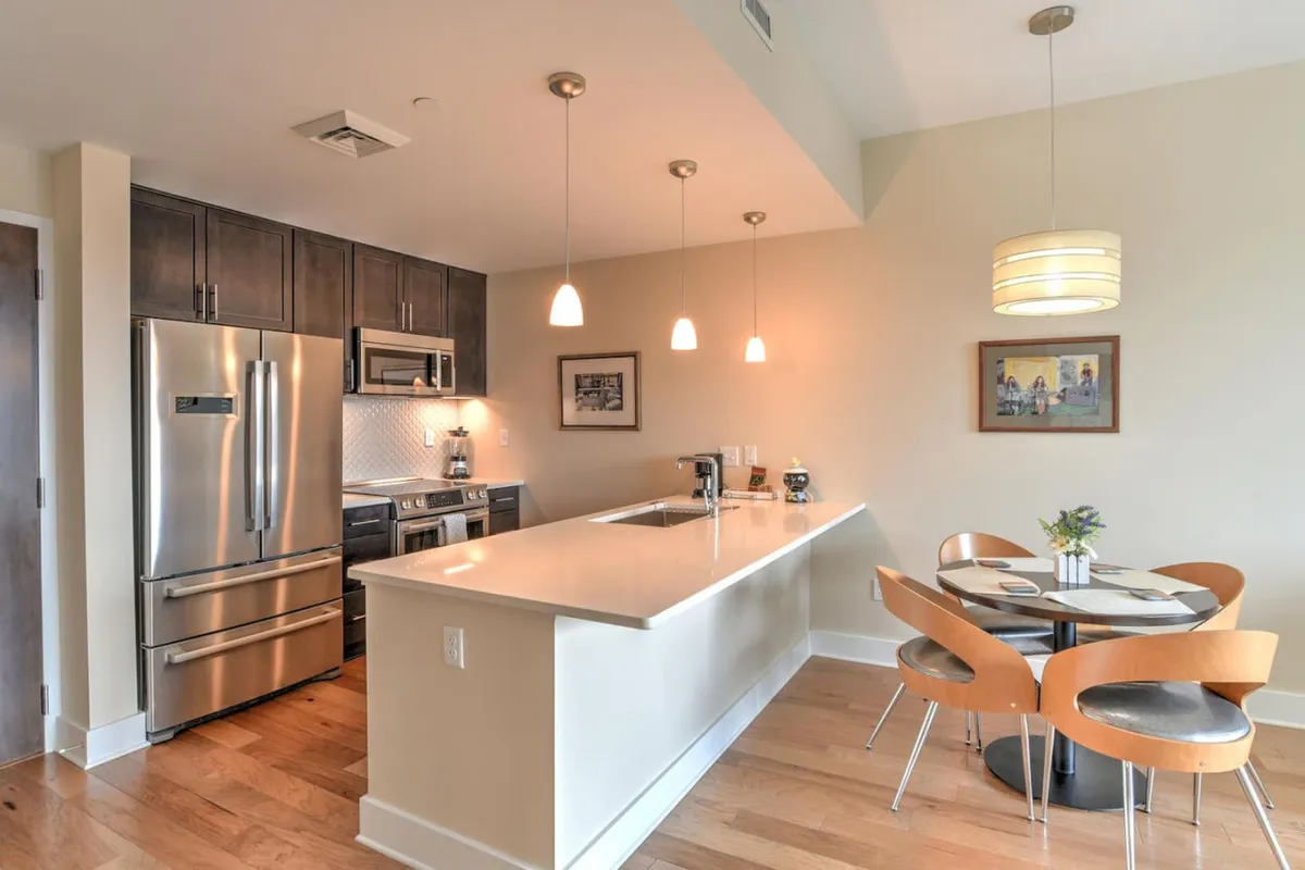 A spacious, modern kitchen featuring a large island with stools, sleek kitchen appliances, pendant lighting, and an adjacent dining table. The inviting setup hints at both professional meal preparation and casual coffee moments, ideal for solo travelers or business guests seeking flexibility.