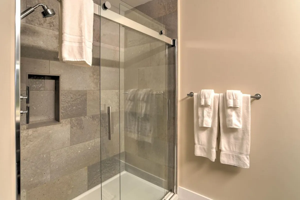 A modern bathroom features a glass-enclosed shower, a sleek tiled wall, and neatly arranged towels on a rack. The space is filled with natural light, emphasizing cleanliness and freshness.