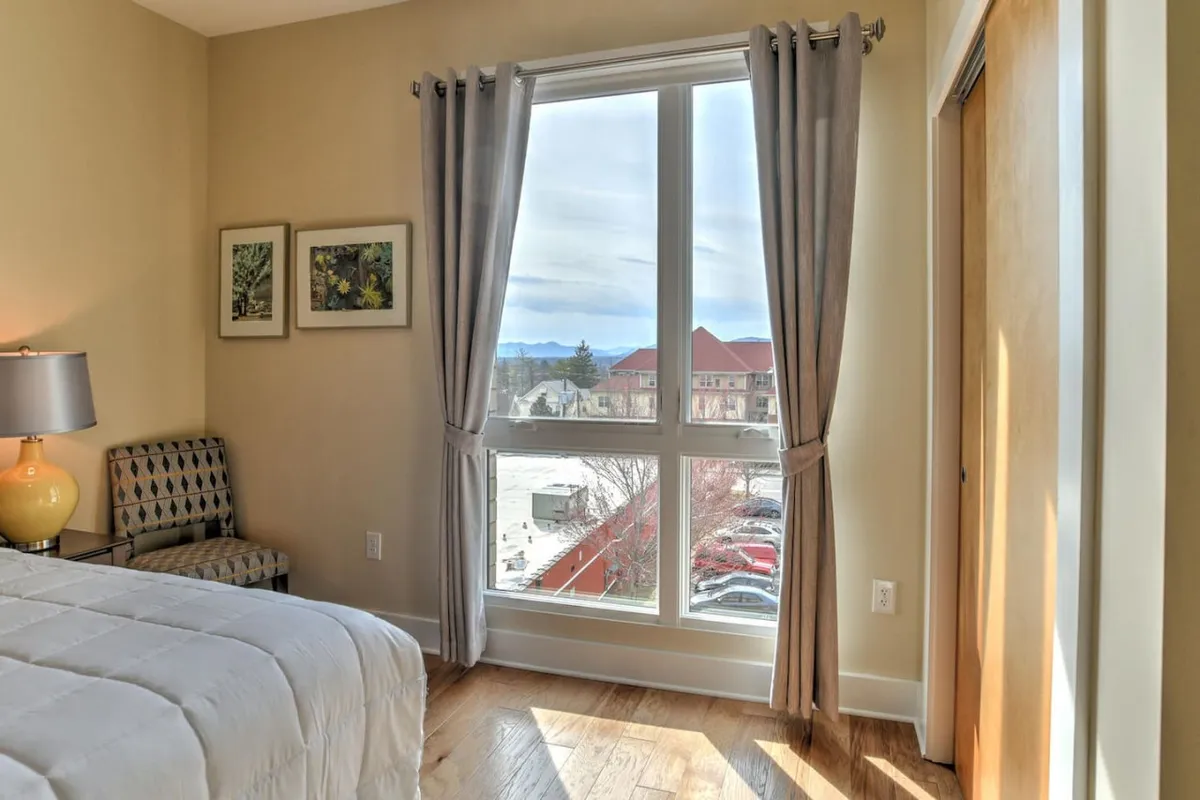A sunlit bedroom features a large window showcasing a mountain view, with a cozy bed dressed in a white comforter, an accent chair, and a warm lamp. The inviting space feels calm and restful, with the outside world visible just beyond the glass.