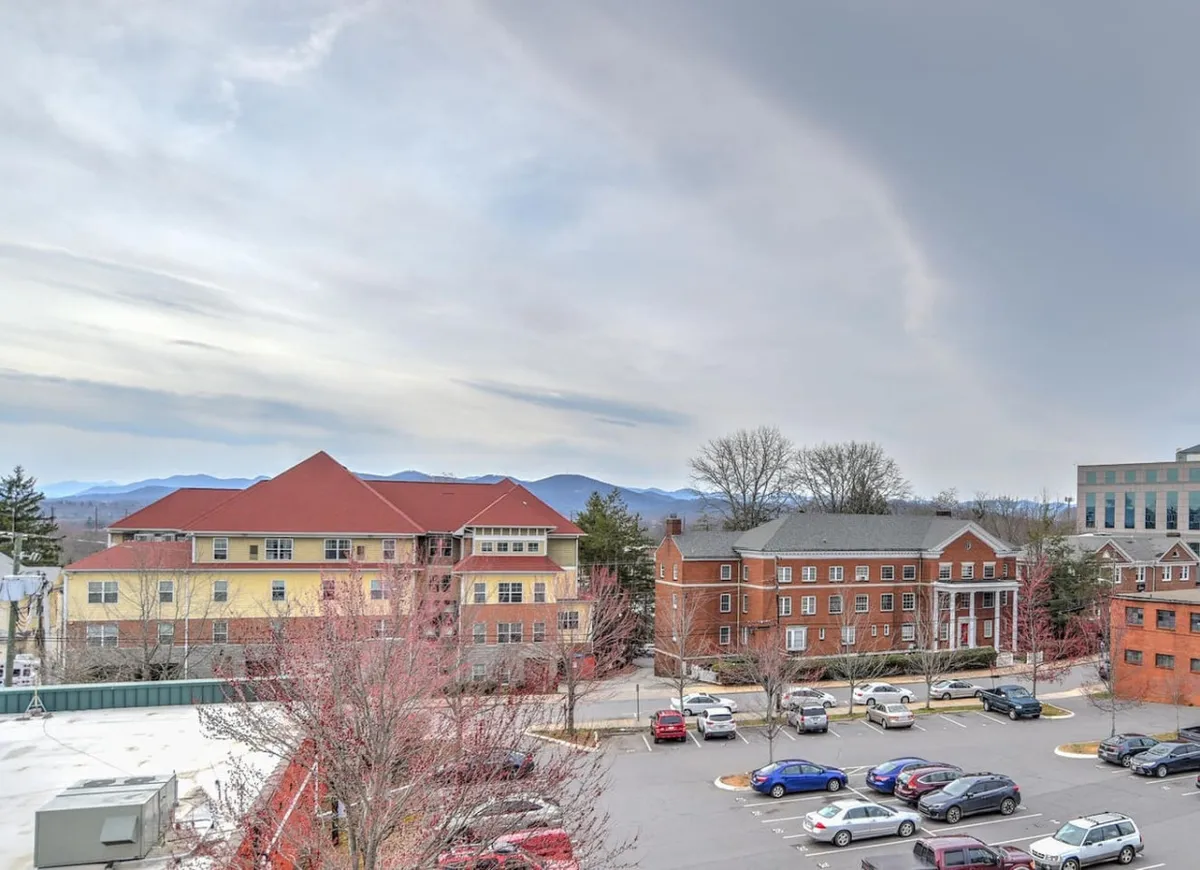 This exterior view highlights the secure building, showing a parking lot in the foreground, with the property surrounded by other buildings and set against a backdrop of mountains and trees. The scene suggests a welcoming, well-maintained communal environment and clearly displays available parking and the overall neighborhood feel.