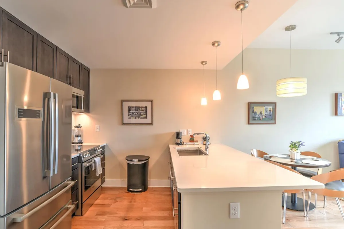 A modern kitchen and dining area featuring stainless steel appliances, a pristine white countertop, and pendant lighting. The dining table is positioned to take in the view from large windows, suggesting a seamless transition from hosting friends at home to heading out for a night on the town.